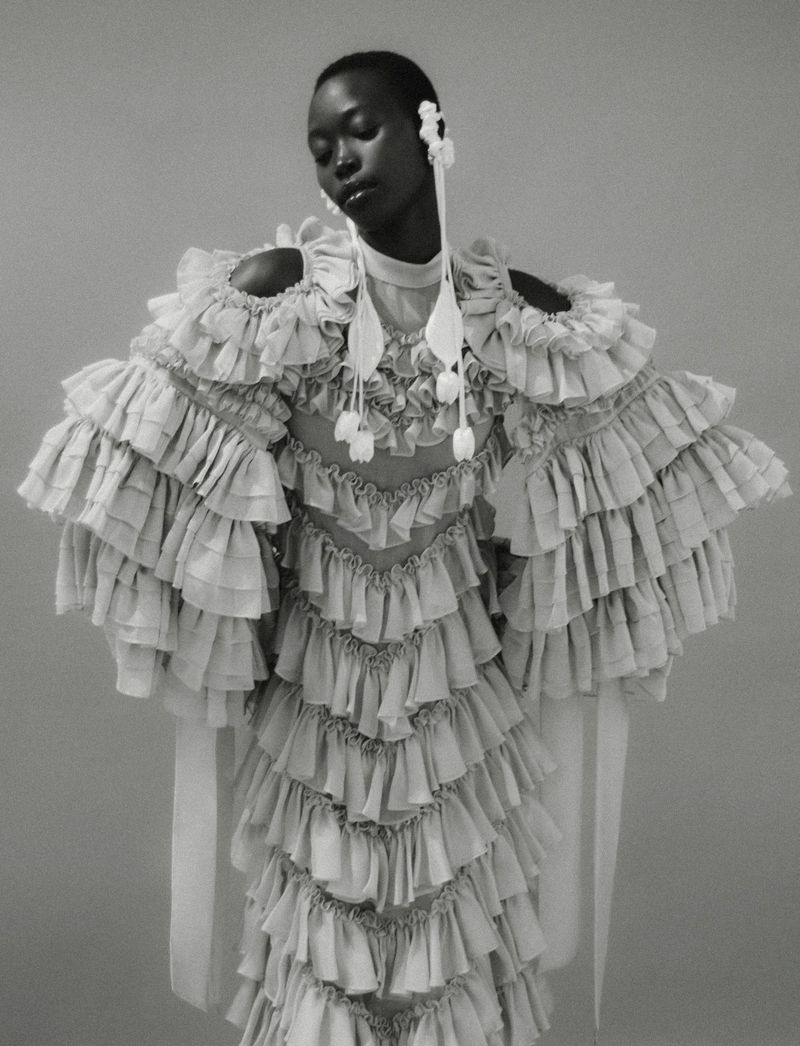 Black woman wearing a sculptural tiered ruffle gown and long tassel earrings in a black and white studio portrait.