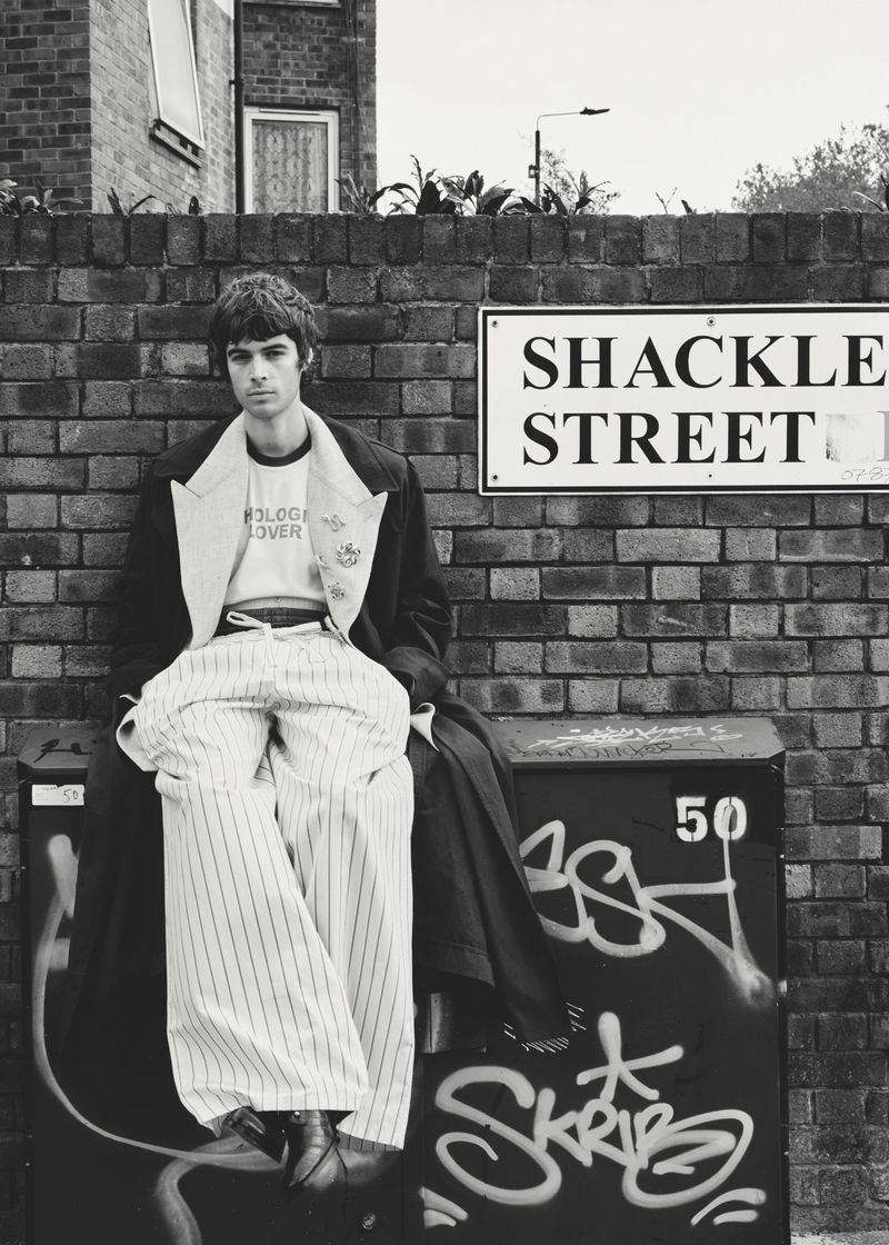 Model Tom Meacock seated on a graffiti-covered utility box wearing wide pinstripe pants before a brick wall and street sign.