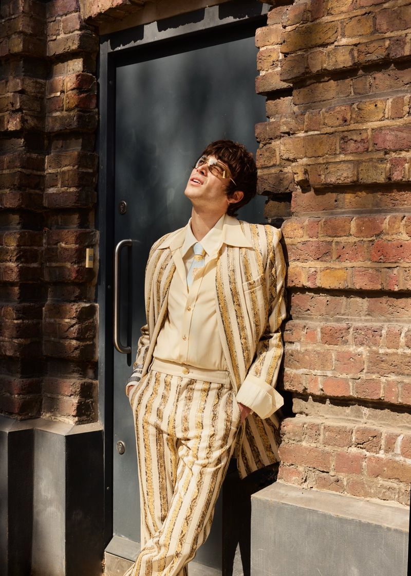A man wearing a cream and gold striped suit, light shirt, and tinted glasses leaning against a dark door in a red brick wall.