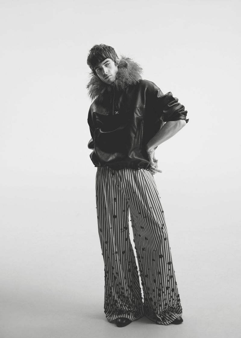 Model Tom Meacock posing in a black fur-trimmed hooded leather jacket and wide-leg striped trousers against a white seamless backdrop.