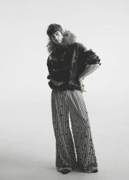 Model Tom Meacock posing in a black fur-trimmed hooded leather jacket and wide-leg striped trousers against a white seamless backdrop.