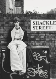 Model Tom Meacock seated on a graffiti-covered utility box wearing wide pinstripe pants before a brick wall and street sign.