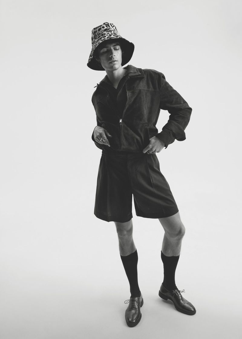 Male model in a patterned bucket hat, dark zip jacket, tailored shorts, high socks, and dress shoes against a white studio background.