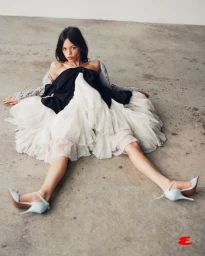 Jenna Ortega sitting on a concrete floor in a black bodice and voluminous tiered white tulle skirt wearing light blue stiletto heels.