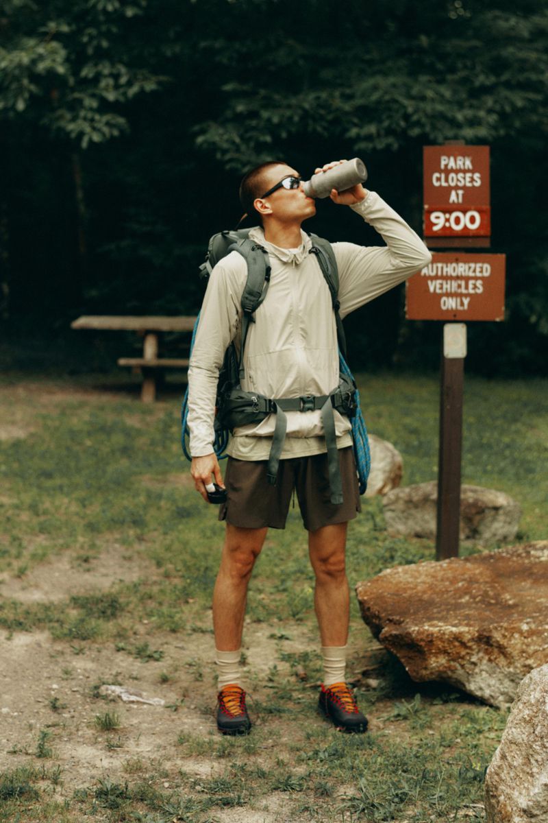A man wearing technical outdoor clothing and a backpack drinks from a water bottle in front of a wooden park sign and trees.