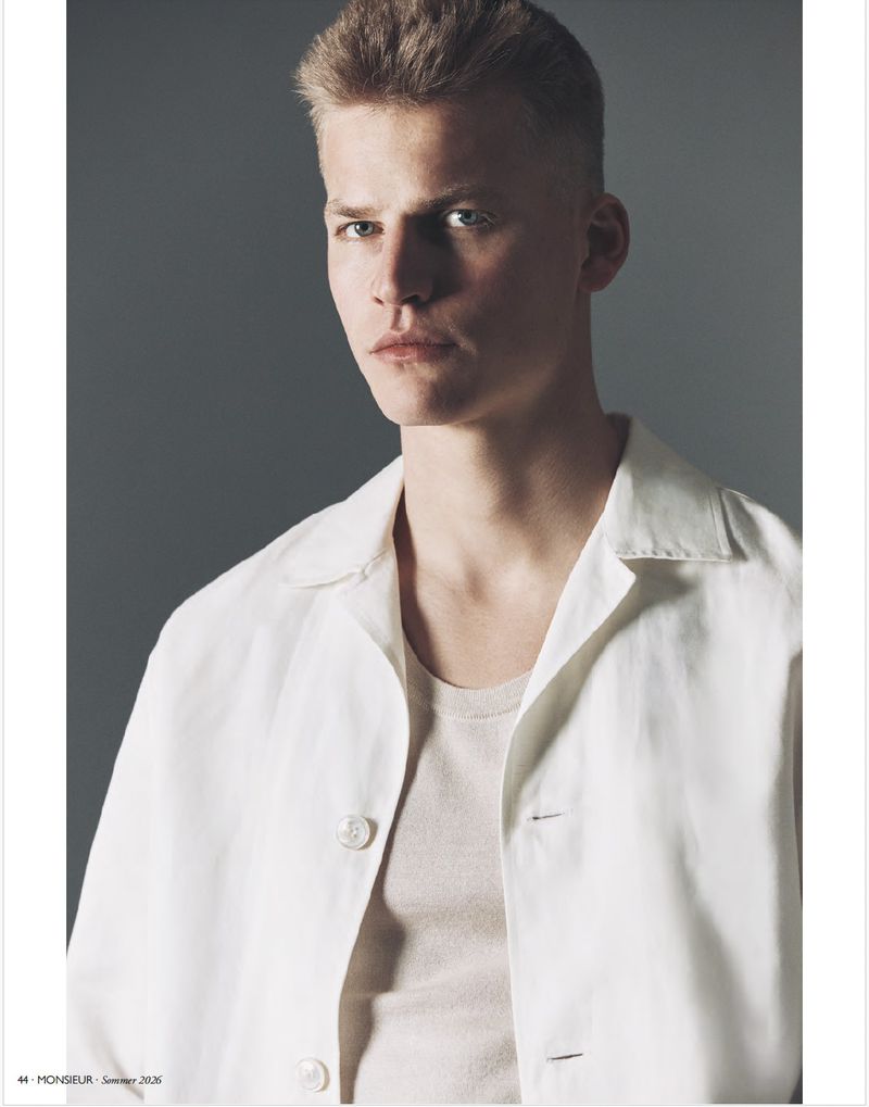 Young man with short blonde hair wearing a white collared shirt over a beige tank top against a plain grey background.