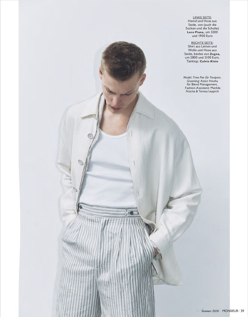 Model Timo Pan looks downward wearing an open off-white overshirt, white tank top, and striped pleated trousers against a white backdrop.