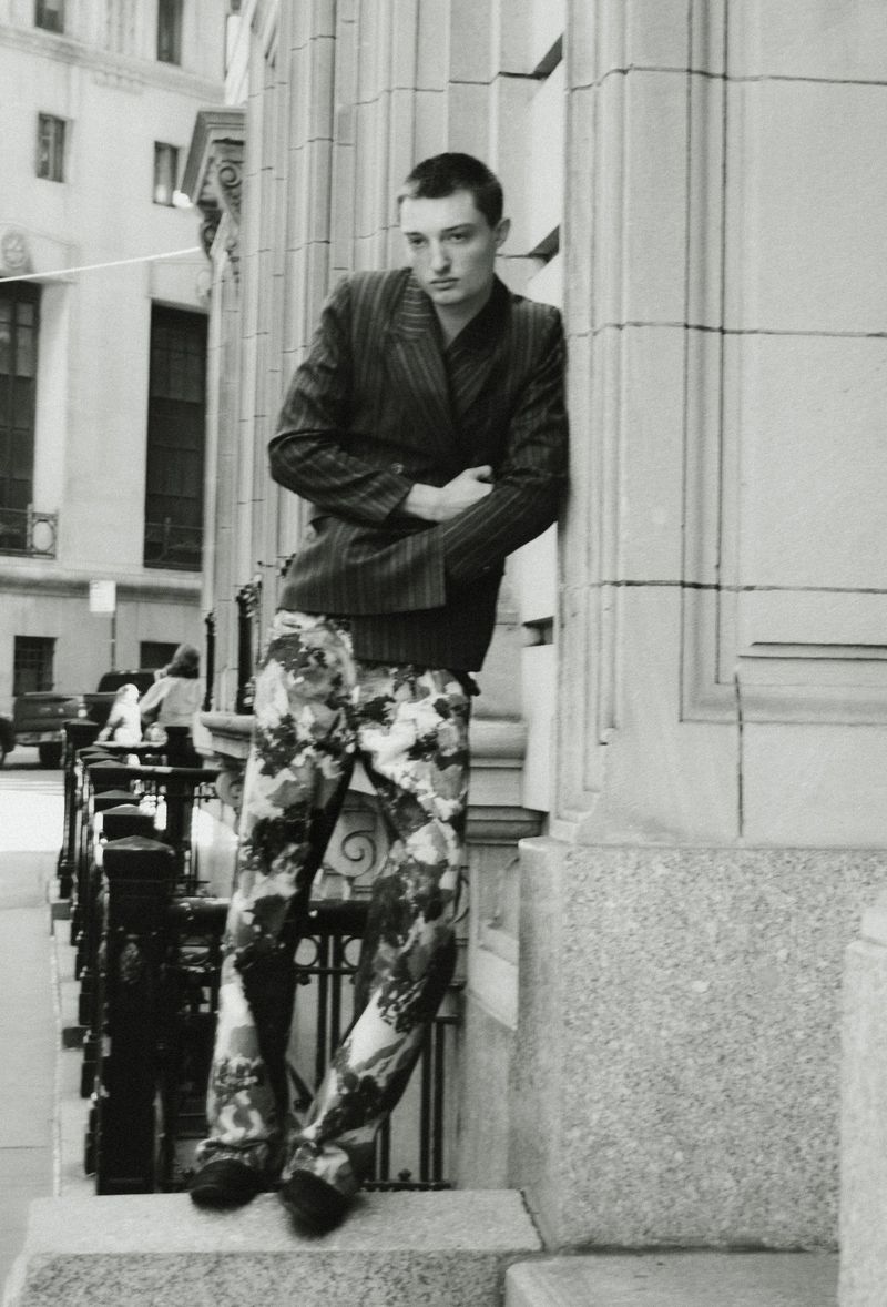 Young man with short hair wearing a pinstripe blazer and patterned trousers leaning against a stone building pillar on a city street.