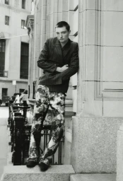 Black and white portrait of a man in a pinstripe blazer and patterned trousers leaning against an exterior stone pillar outdoors.