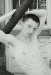 Black and white portrait of a shirtless young man with a buzzcut leaning against a dark wooden beam outdoors.
