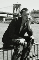 Monochrome portrait of a young man resting his chin on his hand while seated on a railing with the Brooklyn Bridge in the background.