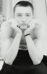 Black and white portrait of a young shirtless man resting his chin on his hands with a direct gaze.