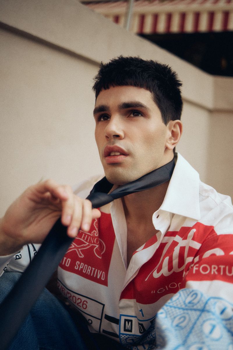 Model Denis Jovanovic pulls a loose black necktie while wearing a red and white graphic collared shirt outdoors.