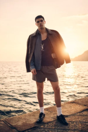 Model Denis Jovanovic stands on a stone pier by the sea wearing layered jackets and shorts during golden hour.