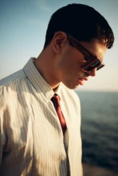 Man wearing chunky brown sunglasses, a striped dress shirt, and a burgundy tie standing against an ocean backdrop in warm sunlight.