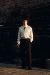 Model Denis Jovanovic in an off-white shirt, dark tie, and brown pleated trousers standing against dark coastal rocks.