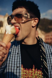 A man wearing oversized wrap sunglasses and a blue plaid shirt eating an ice cream cone outdoors.