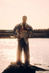 Denis Jovanovic stands on a coastal rock wearing wide-leg trousers and a patterned collared shirt in warm sunlight.