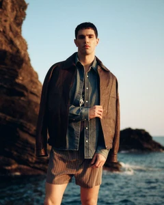 Model Denis Jovanovic wearing a brown leather jacket, denim shirt, and striped shorts against a rocky coast at sunset.