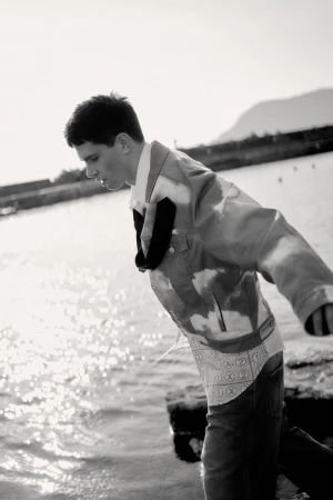 Male model Denis Jovanovic wearing a patterned jacket and collared shirt standing near sparkling coastal water in monochrome.