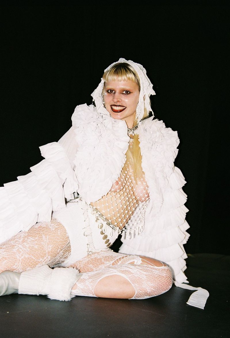 Woman poses sitting indoors in a white ruffled outfit with lace tights against a dark background. Fashion and editorial photography.