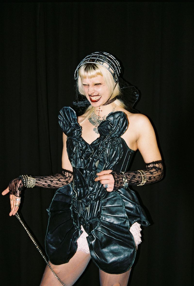 Woman with platinum blonde hair wearing a black ruffled dress, metal headpiece and lace gloves, standing in front of a black backdrop.