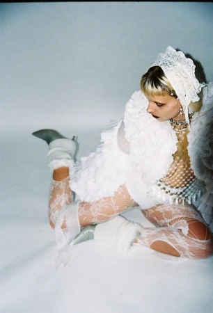 Model in a white lace bonnet, ruffled jacket, and white lace tights reclining on a pale seamless surface.