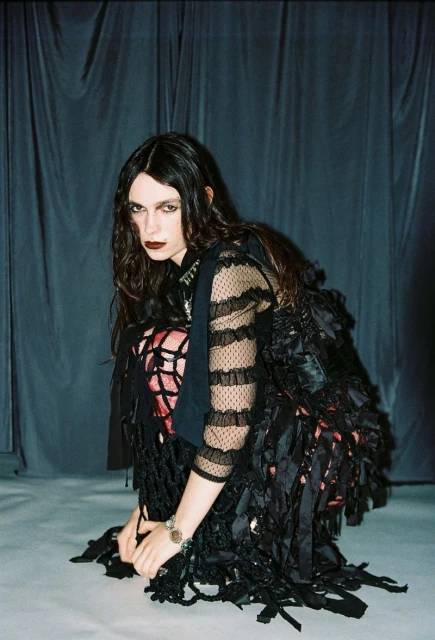 A long-haired woman with dark makeup and a black dress poses crouching on a gray surface against a dark curtain backdrop.