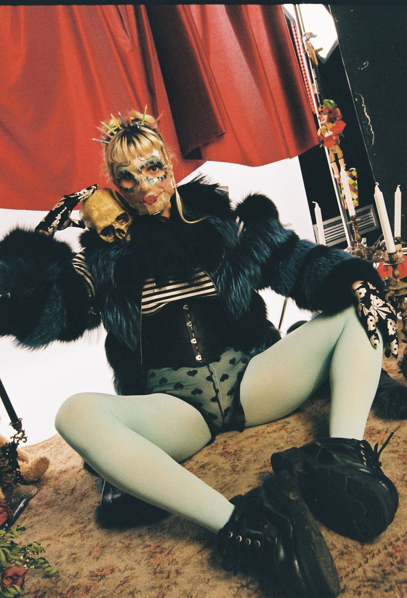 A person with a face mask poses in a fur coat and tights, holding a skull in an indoor environment with props and red background.