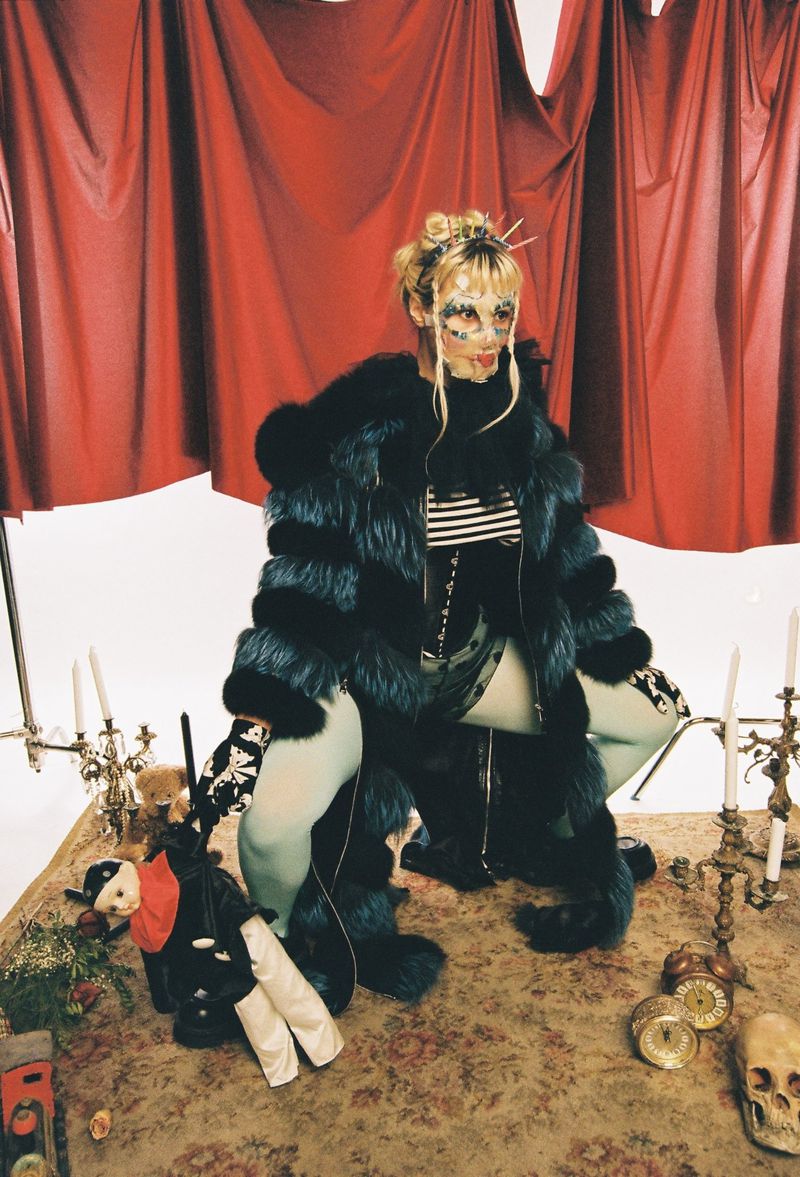 Person in fur coat and decorative mask squats on a floral tablecloth beside dolls, candles, and a skull against a red curtain backdrop.