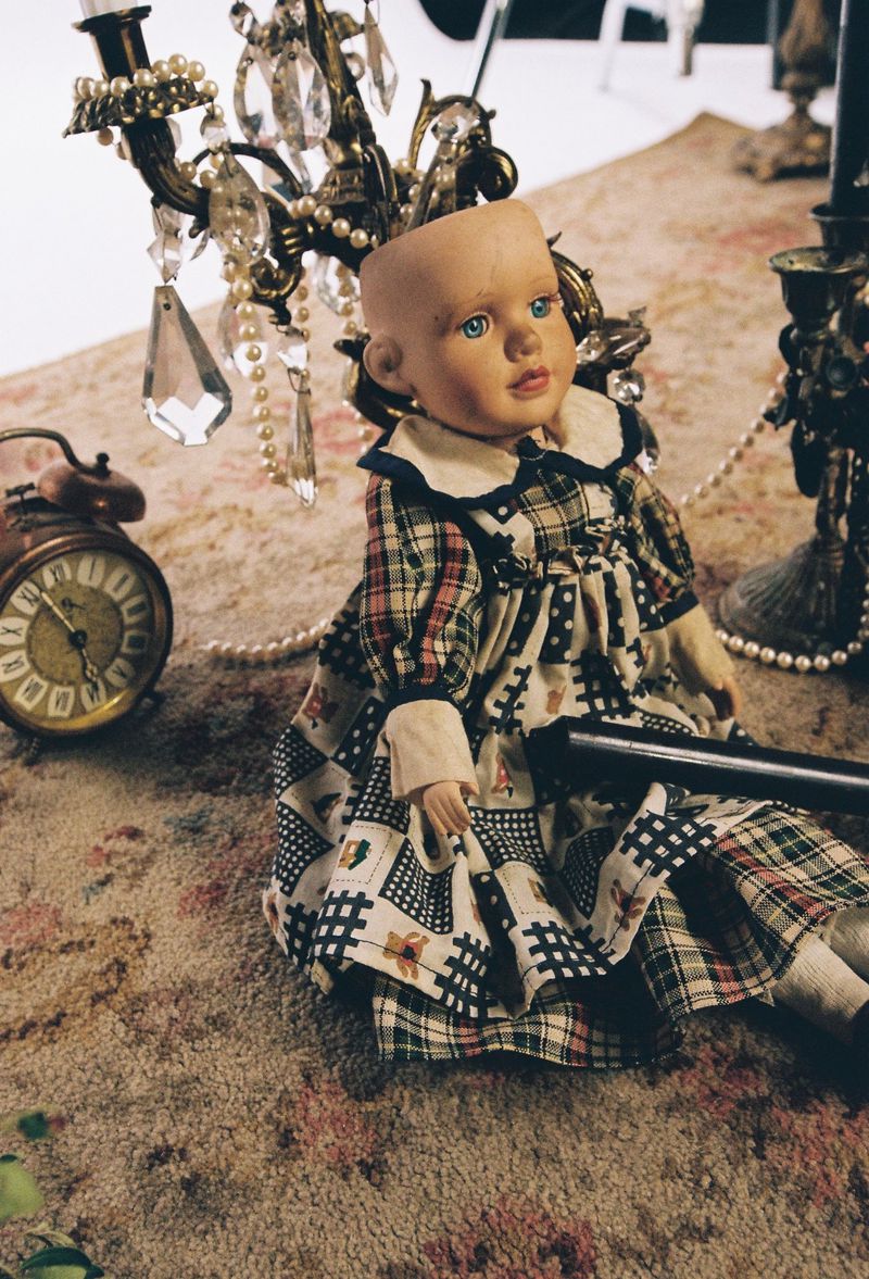 Antique doll in patterned dress sits on a carpet near a vintage clock and chandelier with crystal pendants and pearl strands.