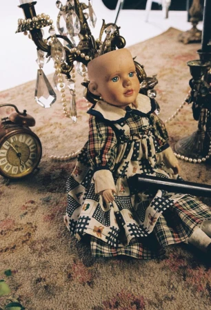Antique doll in patterned dress sits on a carpet near a vintage clock and chandelier with crystal pendants and pearl strands.
