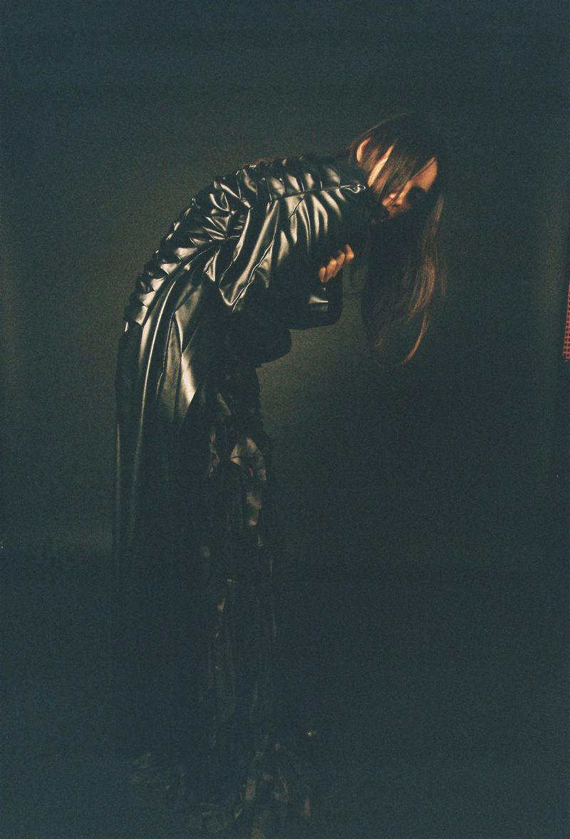 Monochromatic portrait of a person in a long black coat, head bowed, in a dimly lit indoor studio environment with dark shadows.