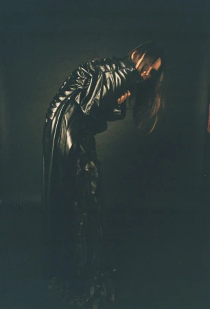 Monochromatic portrait of a person in a long black coat, head bowed, in a dimly lit indoor studio environment with dark shadows.