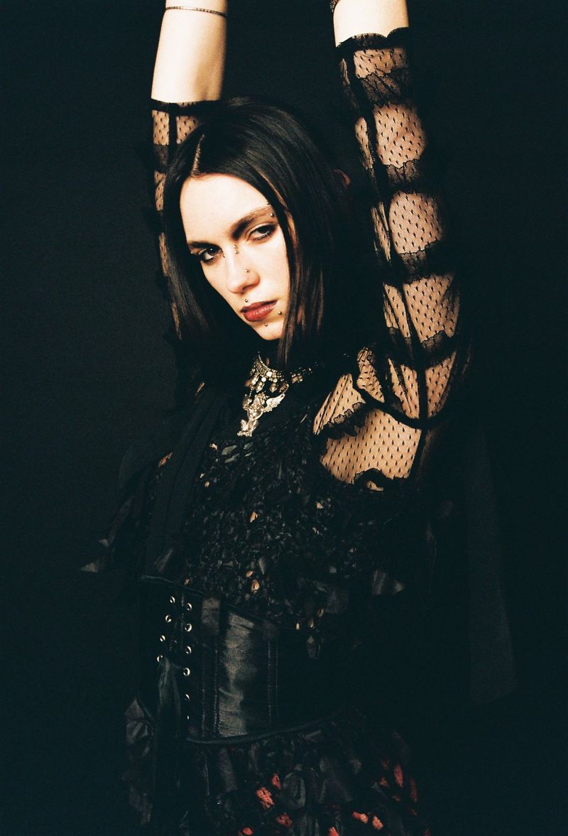 A pale woman with dark hair stands with arms raised, wearing a black lace top and corset against a dark backdrop in a studio shot.