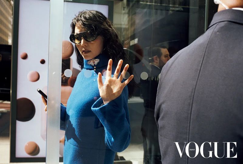A woman wearing a vibrant blue turtleneck and dark sunglasses touches a glass surface while holding a phone, with a man beside her.