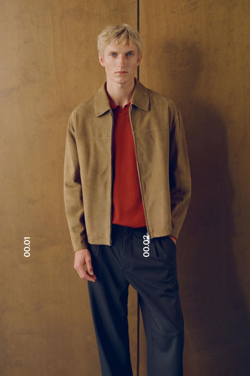Blonde male model wearing a tan suede bomber jacket and red polo shirt with dark navy trousers standing against a wooden wall.