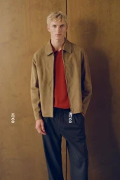 Model Pierrick Grégoire wearing a tan suede jacket, red polo shirt, and dark trousers against a warm textured backdrop.