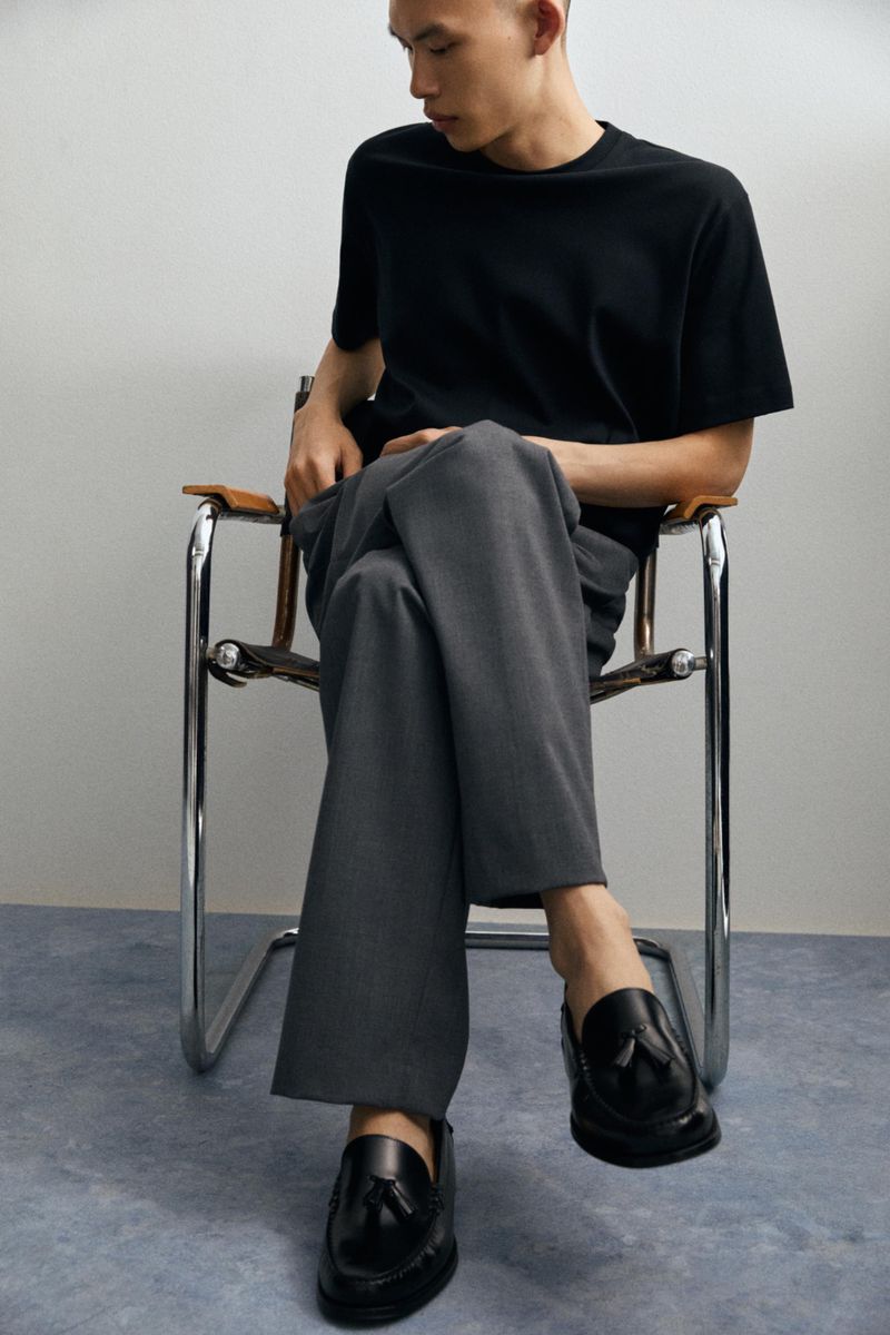 Man wearing a black t-shirt and grey trousers sitting on a chrome-framed leather chair, showcasing black leather tassel loafers.