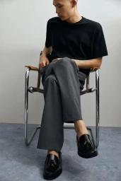 Model Yura Nakano seated in a chrome chair wearing black tassel loafers, grey trousers, and a black t-shirt against a plain studio wall.