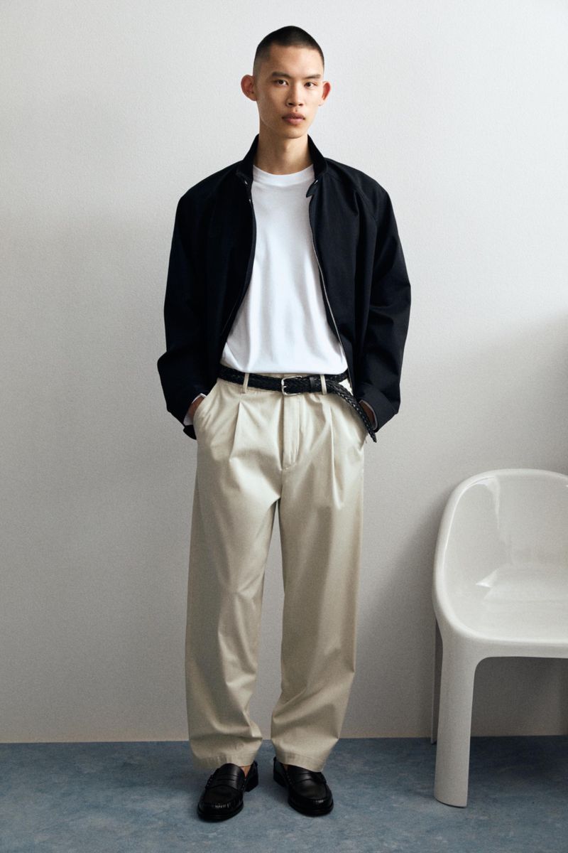 Model Yura Nakano in a black jacket, white t-shirt, beige pleated trousers, and black loafers next to a white chair.