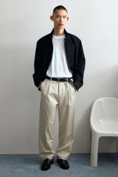 Model Yura Nakano in a black jacket, white t-shirt, beige pleated trousers, and black loafers next to a white chair.