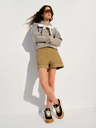 Model Dalene Weber posing in a grey sweatshirt with a white bow, khaki shorts, and oversized sunglasses against a white background.