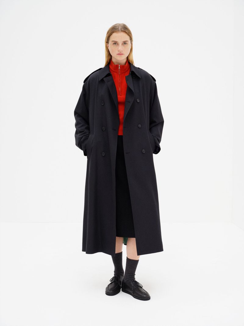 Model Thialda Bok wearing a long black coat over a red zip-up collar with black shoes against a white studio background.