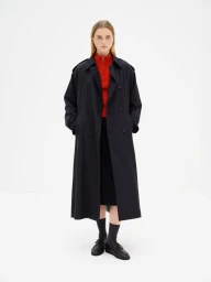 Model Thialda Bok wearing a long black coat over a red zip-up collar with black shoes against a white studio background.