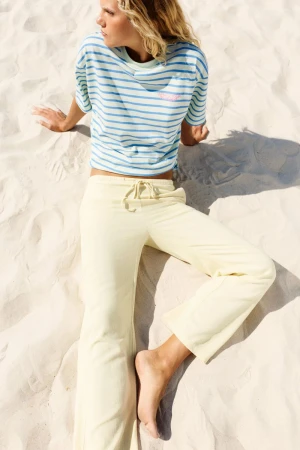 Model Anna Goryainova reclining on white beach sand wearing a blue striped t-shirt and pale yellow drawstring pants.