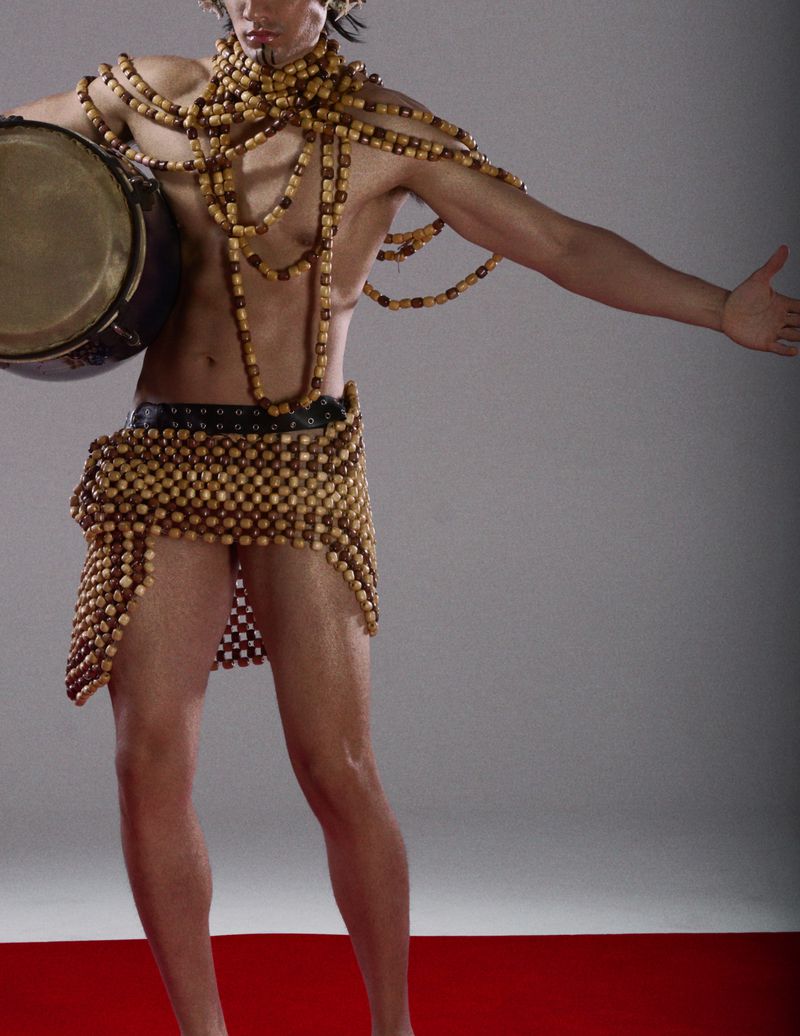 Adult male in a beaded costume poses with a drum on a red surface in a studio, showcasing fashion and artistic expression with dramatic flair.