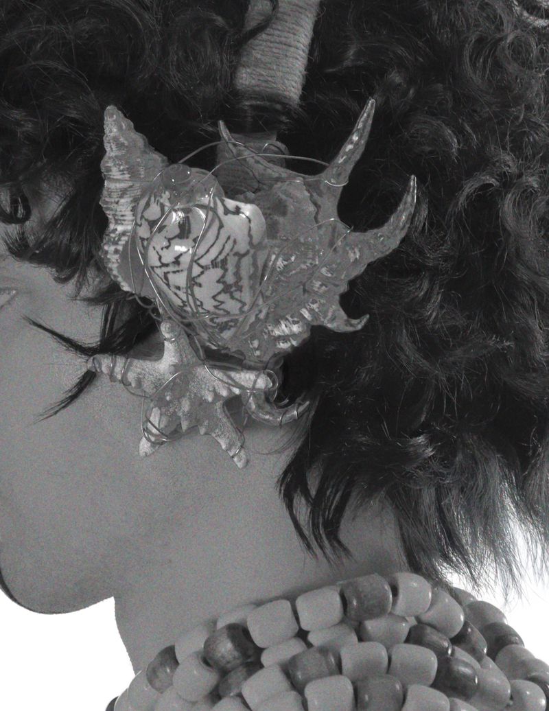 Monochrome close-up of a person with curly hair, wearing a seashell hair accessory and beaded necklace. Black and white fashion shot.