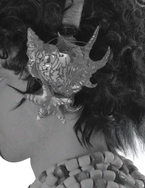 Monochrome close-up of a person with curly hair, wearing a seashell hair accessory and beaded necklace. Black and white fashion shot.