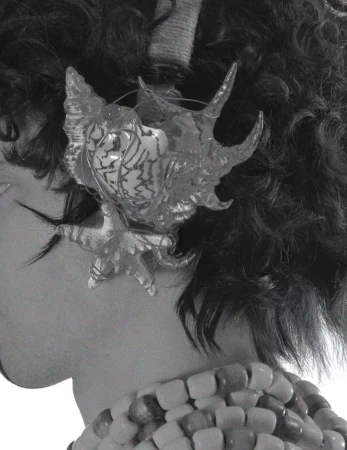 Monochrome close-up of a person with curly hair, wearing a seashell hair accessory and beaded necklace. Black and white fashion shot.